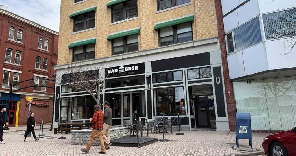 A street view of the Bad Brgr restaurant, with outdoor seating, a few pedestrians walking by, and buildings surrounding the eatery on a cloudy day.