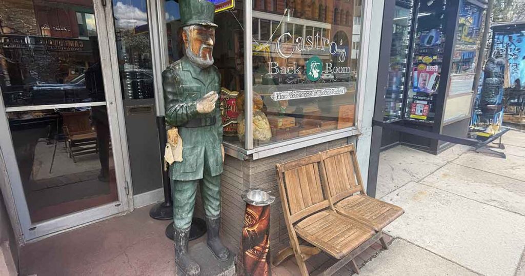 A wooden statue of a bearded man in a green uniform stands outside a storefront near two wooden chairs and a trash can. The window displays signs for a back room and a craft beer establishment.