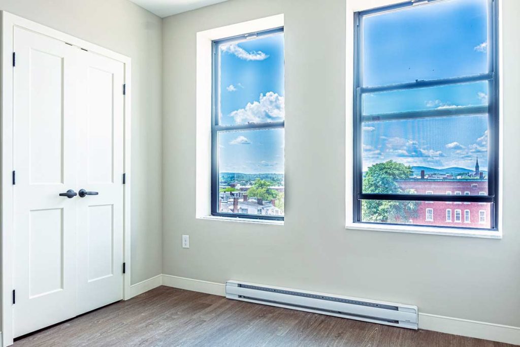 Bright, empty room with light-colored walls, two tall windows showing a cityscape and blue sky, white double doors, wood floor, and a baseboard heater.