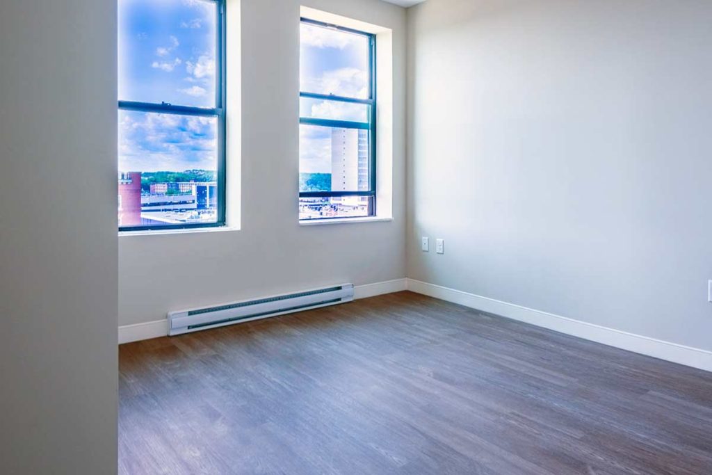 Empty room with light-colored walls, two large windows showing a city view, baseboard heater, and wood-style flooring. Bright natural light fills the minimalist space.