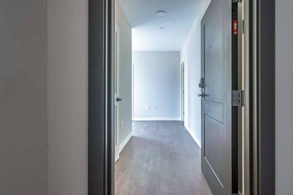 A view from inside an open doorway reveals a hallway with gray walls, wood flooring, and bright natural light coming from an open room at the end.