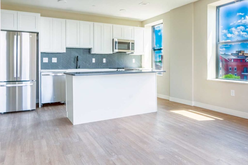 Bright modern kitchen with white cabinets, stainless steel appliances, a central island, wood flooring, and large windows letting in natural light and showing a city view with blue sky.