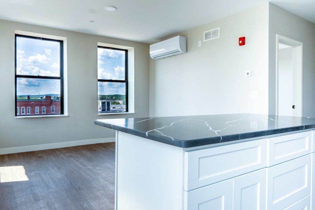 Bright, modern apartment interior with large windows, white walls, hardwood floors, and a black countertop kitchen island. An air conditioning unit is mounted on the wall above.