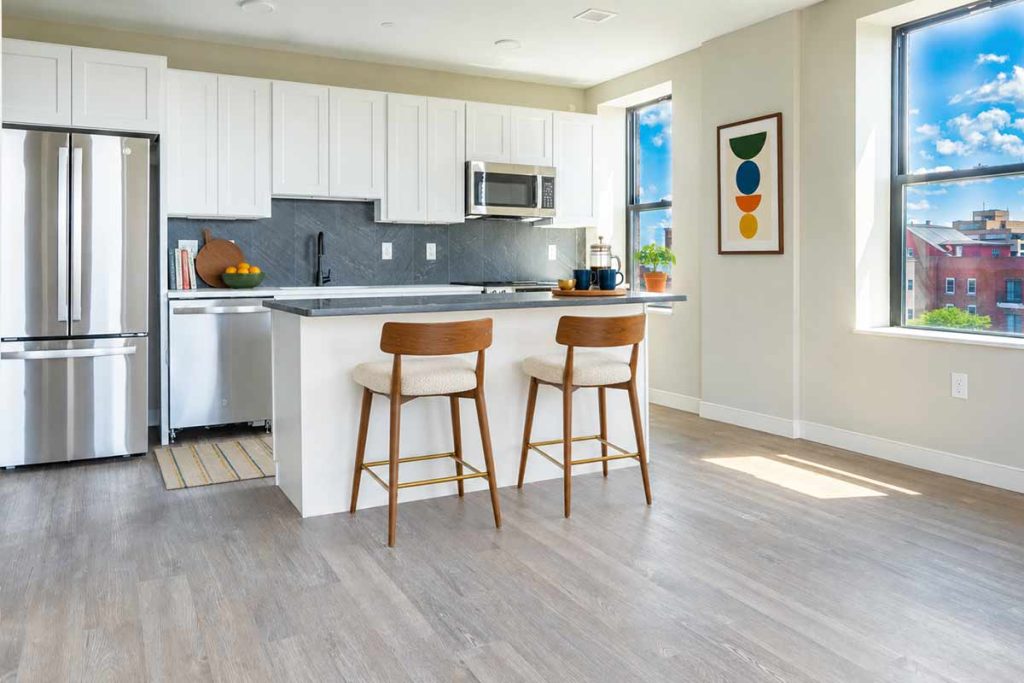 Modern kitchen with white cabinets, stainless steel appliances, island with two wooden barstools, a framed abstract art piece, potted plant, and large sunny windows with a view of buildings outside.