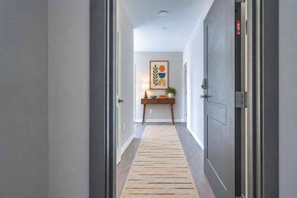 A modern hallway with a striped rug, a wooden console table holding a lamp, bowl, and plant, and abstract art hanging above the table. The space has light walls and dark doors.