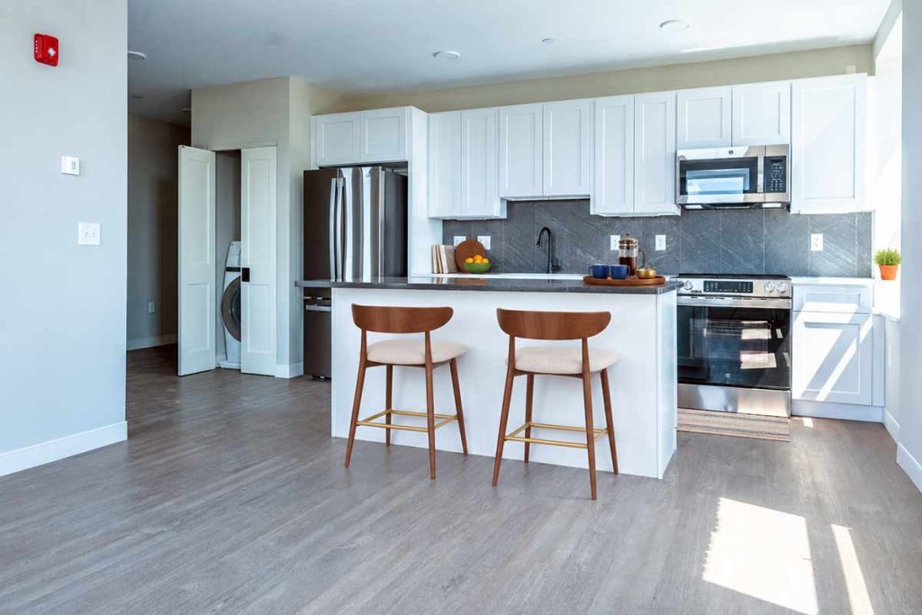 Modern kitchen with white cabinets, stainless steel appliances, a gray backsplash, and a central island with two wooden chairs. Sunlight streams in from the right, illuminating the space.
