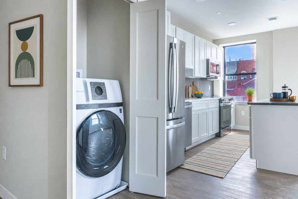 A modern kitchen with white cabinets and stainless steel appliances, next to a laundry area with a front-loading washer partially behind a sliding door. A colorful rug and wall art are visible.