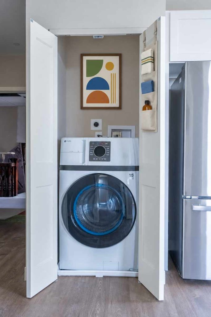 A front-load washing machine sits in a closet with open doors. Above it is modern abstract art. A hanging organizer with small items is on the right door, near a stainless steel refrigerator.